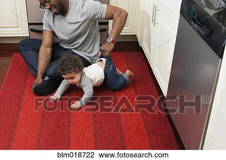 Stock Image - Father helping baby crawl in kitchen. Fotosearch