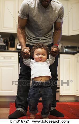 Stock Image - Father helping baby walk in kitchen. Fotosearch