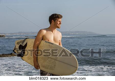 Stock Image - Man carrying surfboard on beach. Fotosearch