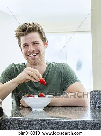 Stock Image - Man eating strawberries in kitchen. Fotosearch