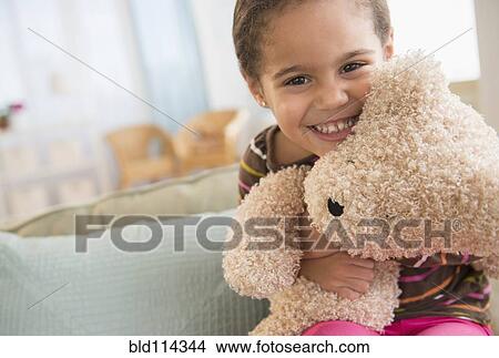 Picture - Hispanic girl hugging teddy bear on sofa. Fotosearch