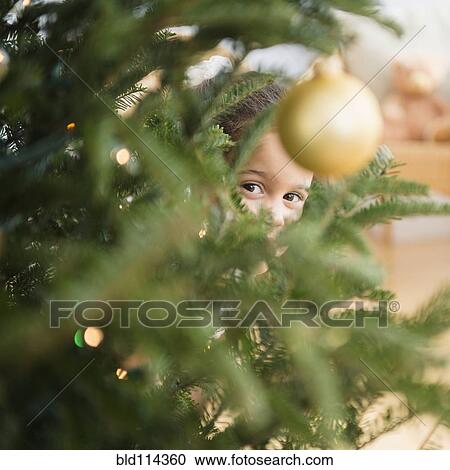 Stock Image - Hispanic girl peeking behind Christmas tree. Fotosearch