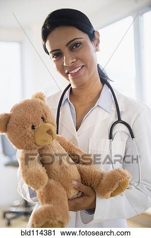 Stock Image - Indian doctor holding teddy bear in office. Fotosearch
