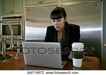 Stock Image - Mixed race businesswoman using laptop in kitchen. Fotosearch