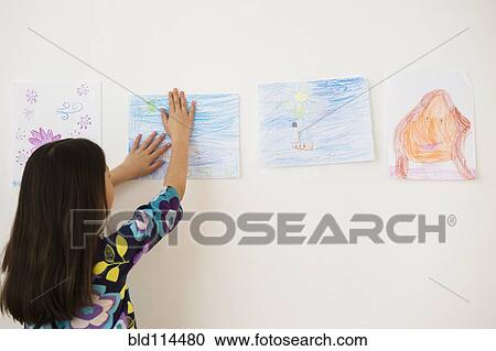 Stock Image - Mixed race girl hanging drawings on wall. Fotosearch