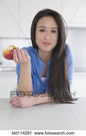 Stock Image - Mixed race woman eating fruit in kitchen. Fotosearch