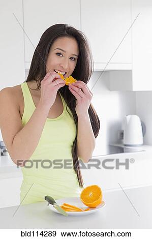 Stock Photo - Mixed race woman eating fruit in kitchen. Fotosearch