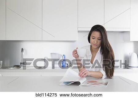 Stock Photography - Mixed race woman reading magazine in kitchen. Fotosearch