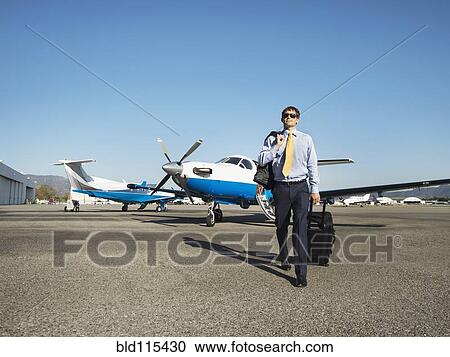 Stock Image - Caucasian businessman rolling luggage on runway. Fotosearch