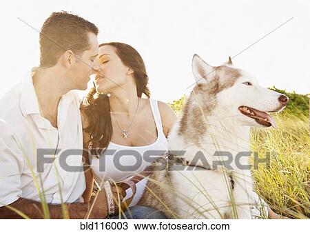 Stock Image - Caucasian couple sitting with dog in tall grass. Fotosearch