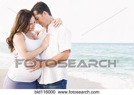 Stock Image - Caucasian man hugging pregnant wife on beach. Fotosearch