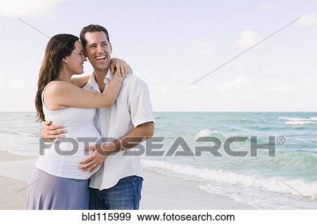 Stock Photo - Caucasian man hugging pregnant wife on beach. Fotosearch