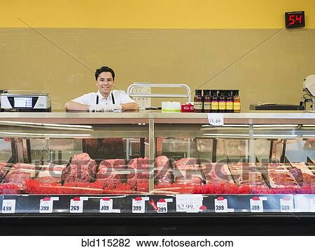 Stock Image - Hispanic butcher at meat counter of grocery store. Fotosearch