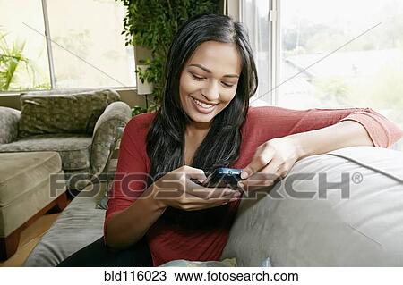 Stock Image - Indian woman using cell phone on sofa. Fotosearch