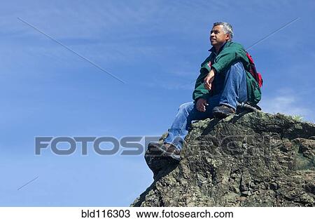 Stock Image - Mixed race man sitting on rocky hilltop. Fotosearch