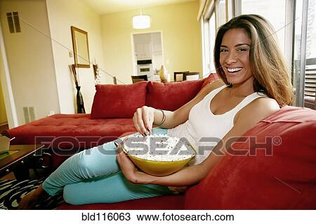 Stock Image - Mixed race woman eating popcorn on sofa. Fotosearch