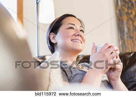 Stock Image - Mixed race woman having cup of coffee on sofa. Fotosearch
