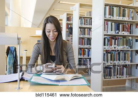 Stock Image - Student using cell phone at desk in library. Fotosearch