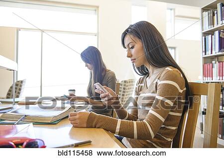 Stock Photo - Student using cell phone at desk in library. Fotosearch