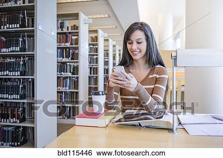 Stock Photograph - Student using cell phone at desk in library. Fotosearch