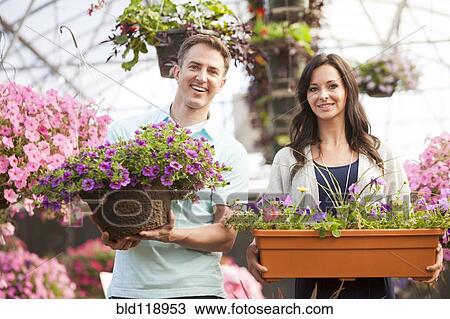 Stock Image - Caucasian couple carrying potted flowers in plant nursery. Fotosearch