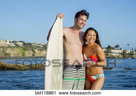 Stock Image - Couple with surfboard smiling together on beach. Fotosearch