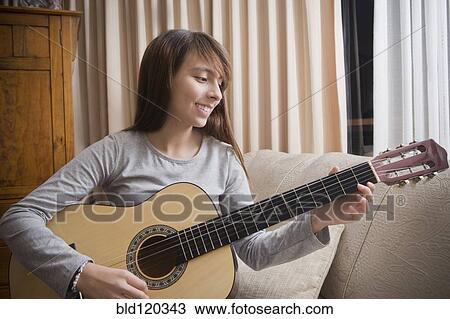 Stock Image - Hispanic girl playing guitar on sofa. Fotosearch