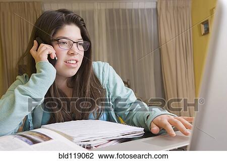 Stock Image - Hispanic girl talking on cell phone at laptop. Fotosearch