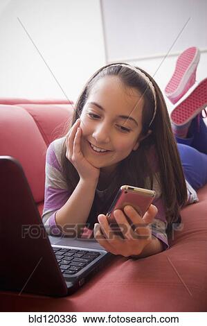 Stock Photograph - Hispanic girl using laptop and cell phone on sofa. Fotosearch
