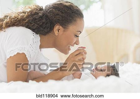 Picture - Hispanic mother playing with infant on bed. Fotosearch