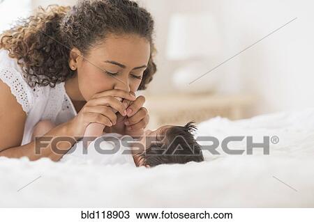 Stock Image - Hispanic mother playing with infant on bed. Fotosearch