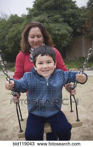 Stock Image - Hispanic mother pushing son on swings. Fotosearch