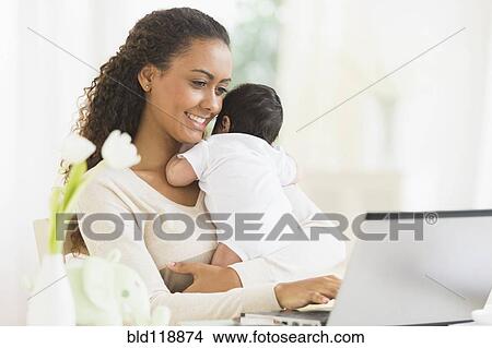 Picture - Hispanic mother using laptop and holding infant son. Fotosearch