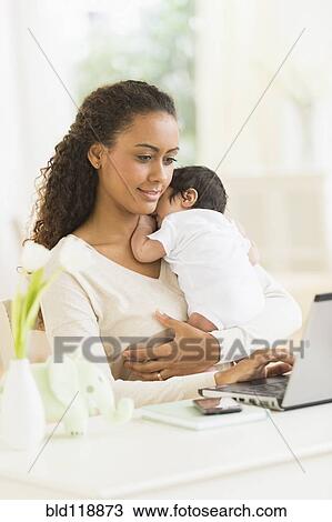 Stock Image - Hispanic mother using laptop and holding infant son. Fotosearch