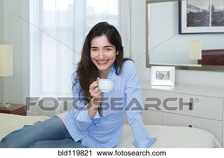 Stock Image - Hispanic woman enjoying cup of coffee on bed. Fotosearch