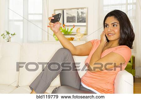 Stock Photo - Hispanic woman taking self-portrait with camera phone on sofa. Fotosearch