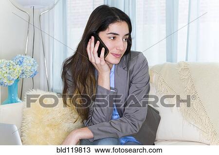 Stock Image - Hispanic woman talking on cell phone on sofa. Fotosearch
