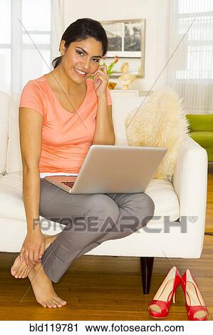 Stock Image - Hispanic woman using laptop and cell phone on sofa. Fotosearch