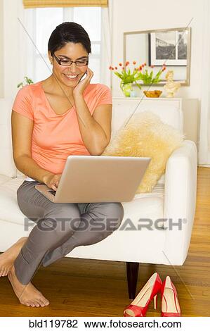 Stock Image - Hispanic woman using laptop and cell phone on sofa. Fotosearch