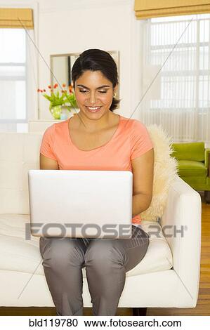 Stock Image - Hispanic woman using laptop on sofa. Fotosearch