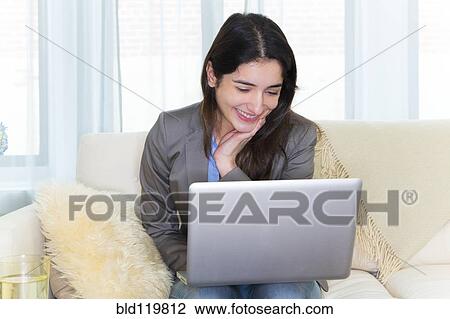 Stock Image - Hispanic woman using laptop on sofa. Fotosearch