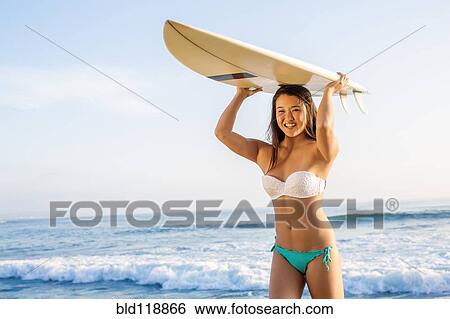 Stock Photograph - Japanese woman carrying surfboard overhead on beach. Fotosearch