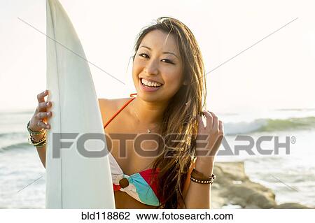 Stock Image - Mixed race woman holding surfboard on beach. Fotosearch