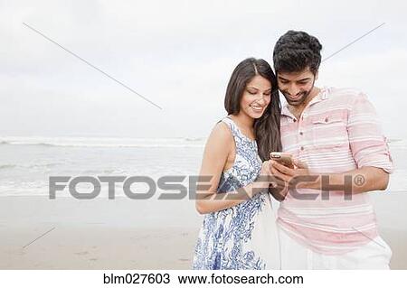 Stock Image - Indian couple using cell phone on beach. Fotosearch