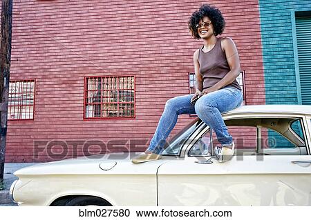 Stock Image - Mixed race woman sitting on vintage car. Fotosearch