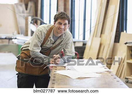 Stock Photo - Caucasian carpenter looking at plan in workshop. Fotosearch