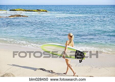 Stock Image - Caucasian woman carrying surfboard on beach. Fotosearch