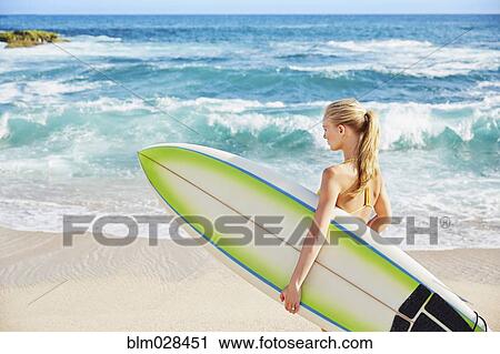 Stock Image - Caucasian woman carrying surfboard on beach. Fotosearch