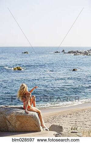 Stock Photo - Caucasian woman sitting on rock on beach. Fotosearch