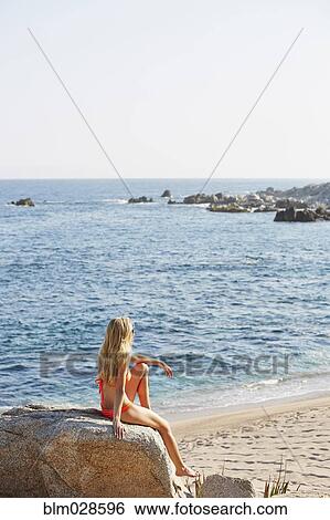 Stock Photograph - Caucasian woman sitting on rock on beach. Fotosearch